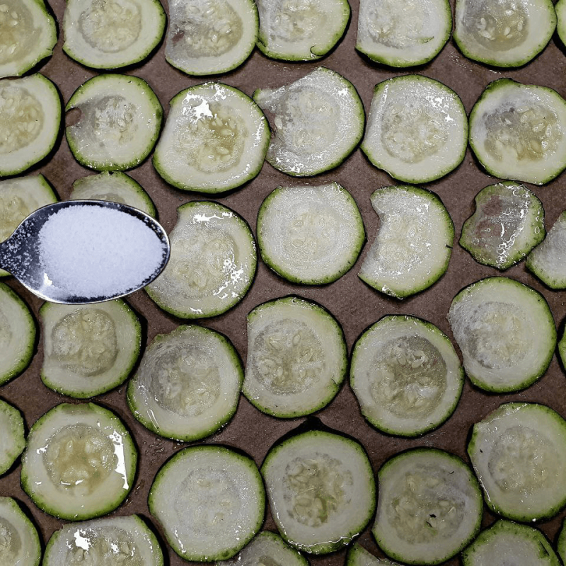 Man sieht einen Löffel mit Salz über Zucchinischeiben, die auf einem Backblech aufgereiht sind und später mal Zucchini Chips werden sollen